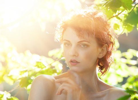 Portrait of a young red-haired woman among the foliage of trees illuminated by the suns rays
