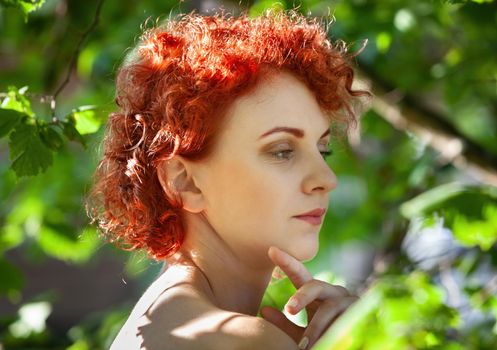 Portrait of a young red-haired woman among the foliage of trees illuminated by the suns rays