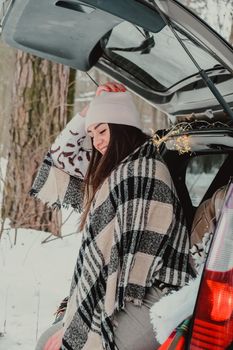 Brunette woman wrapped in blanket in trunk car drinking coffee tea from thermos. Travel in winter. Car decorated with festive Christmas lights. Outdoor picnic. Unity with nature