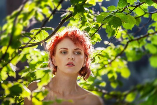 Portrait of a young red-haired woman among the foliage of trees illuminated by the suns rays