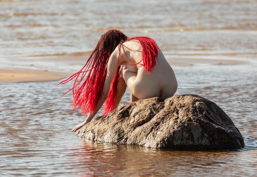 Beautiful girl outdoors enjoying nature. Young naked woman with scarlet dreadlocks on the stone in the sea enjoys the tranquility of nature