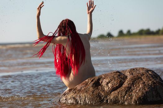 Beautiful girl outdoors enjoying nature. Young naked woman with scarlet dreadlocks on the stone in the sea enjoys the tranquility of nature