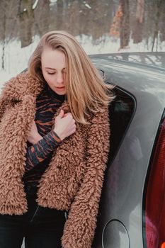 Attractive beautiful young woman in sweater staying next to the car in forest. Winter vacation. Holidays. Smilling Christmas lights.