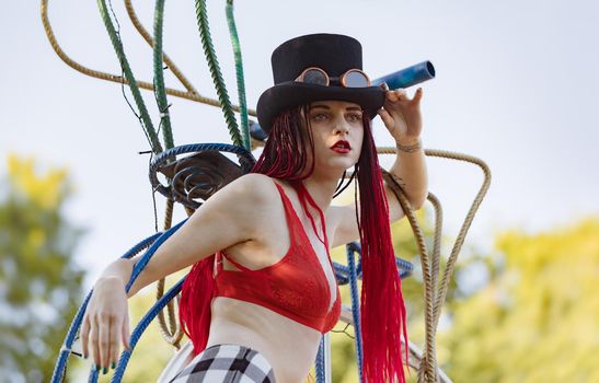 Glamorous girl with scarlet dreadlocks, a red swimsuit, black hat and welding glasses poses on the blue sky background. Free space for your text