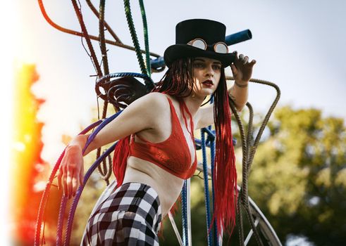 Glamorous girl with scarlet dreadlocks, a red swimsuit, black hat and welding glasses poses on the blue sky background. Free space for your text