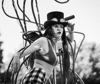 Glamorous girl with scarlet dreadlocks, a red swimsuit, black hat and welding glasses poses on the blue sky background. Free space for your text. Black and white image