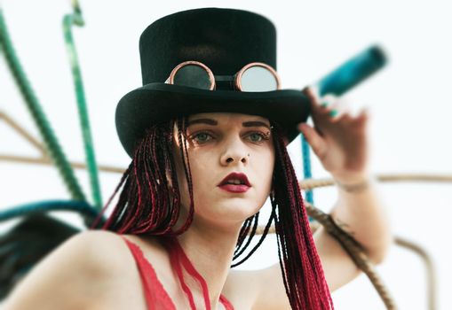 Glamorous girl with scarlet dreadlocks, a red swimsuit, black hat and welding glasses poses on the blue sky background. Free space for your text