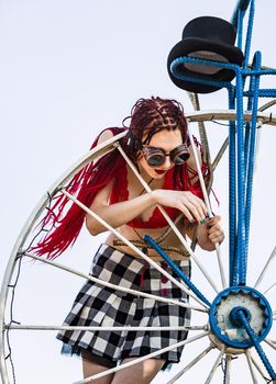 Glamorous girl with scarlet dreadlocks, a red swimsuit, black hat and welding glasses poses on the blue sky background. Free space for your text