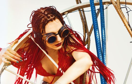 Glamorous girl with scarlet dreadlocks, a red swimsuit, black hat and welding glasses poses on the blue sky background. Free space for your text