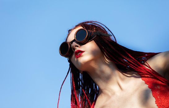 Glamorous girl with scarlet dreadlocks, a red swimsuit and welding glasses poses on the blue sky background. Free space for your text