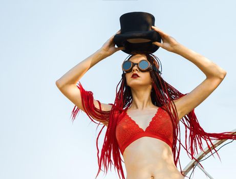 Glamorous girl with scarlet dreadlocks, a red swimsuit, black hat and welding glasses poses on the blue sky background. Free space for your text