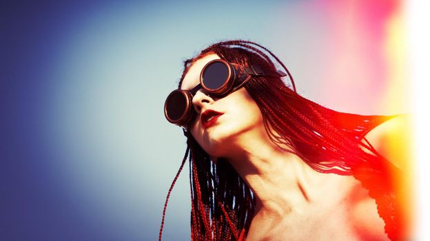 Glamorous girl with scarlet dreadlocks, a red swimsuit and welding glasses poses on the blue sky background. Free space for your text