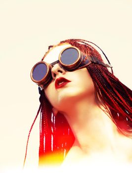 Glamorous girl with scarlet dreadlocks, a red swimsuit and welding glasses poses on the blue sky background. Free space for your text