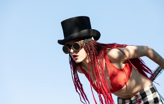 Glamorous girl with scarlet dreadlocks, a red swimsuit, black hat and welding glasses poses on the blue sky background. Free space for your text