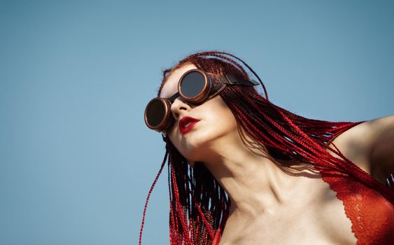 Glamorous girl with scarlet dreadlocks, a red swimsuit and welding glasses poses on the blue sky background. Free space for your text