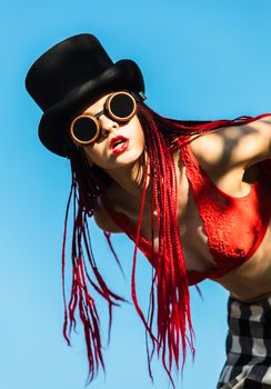 Glamorous girl with scarlet dreadlocks, a red swimsuit, black hat and welding glasses poses on the blue sky background. Free space for your text