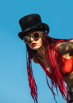 Glamorous girl with scarlet dreadlocks, a red swimsuit, black hat and welding glasses poses on the blue sky background. Free space for your text