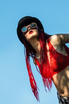 Glamorous girl with scarlet dreadlocks, a red swimsuit, black hat and welding glasses poses on the blue sky background. Free space for your text