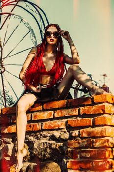 Glamorous girl with scarlet dreadlocks, a red swimsuit and welding glasses sits on a brick wall and poses on the blue sky background. Free space for your text