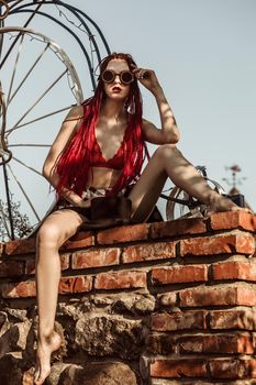 Glamorous girl with scarlet dreadlocks, a red swimsuit and welding glasses sits on a brick wall and poses on the blue sky background. Free space for your text