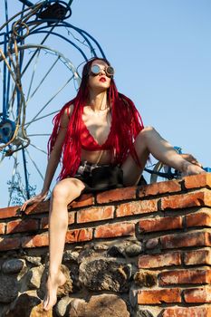 Glamorous girl with scarlet dreadlocks, a red swimsuit and welding glasses sits on a brick wall and poses on the blue sky background. Free space for your text