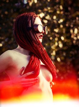 Semi-nude glamorous girl with scarlet dreadlocks and welding glasses closes the breast with scarlet dreadlocks