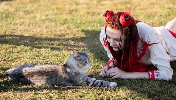 Young woman with scarlet dreadlocks in national dress lying on the grass and playing with the cat. Outdoors portrait