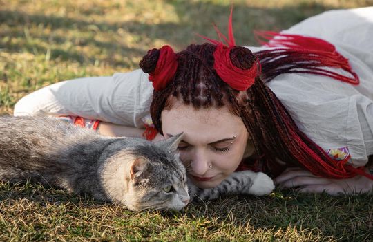 Young woman with scarlet dreadlocks in national dress lying on the grass and playing with the cat. Outdoors portrait