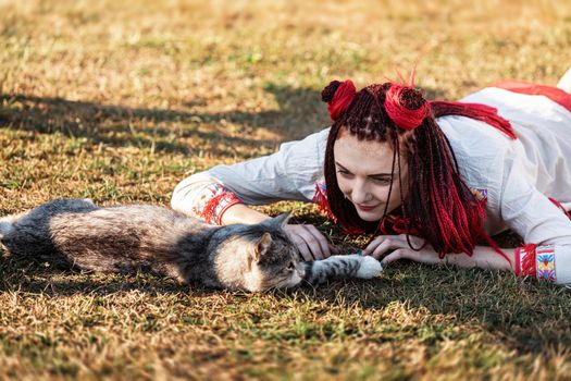 Young woman with scarlet dreadlocks in national dress lying on the grass and playing with the cat. Outdoors portrait
