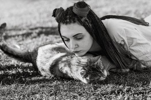 Black and white image of an young woman with dreadlocks in national dress lying on the grass and playing with the cat. Outdoors portrait
