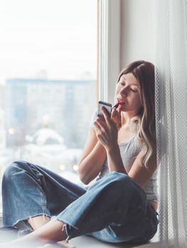 Woman with curly hair paints her lips with lipstick and uses smartphone as mirror. Fast make-up. Morning routine. Cup of hot coffee on windowsill.