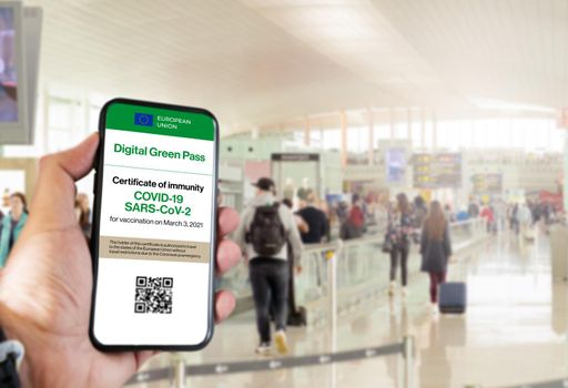 The digital green pass of the european union with the QR code on the screen of a mobile held by a hand with blurred airport in the background. Immunity from Covid-19. Travel without restrictions.