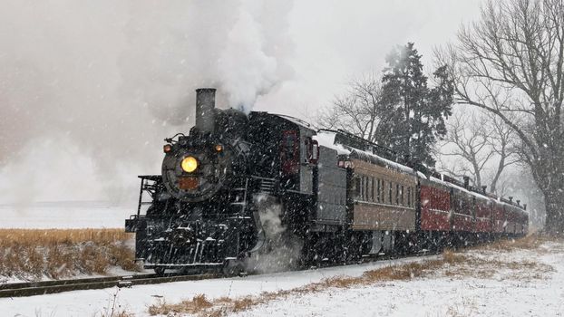 View of An Antique Restored Steam Locomotive Blowing Smoke and Steam Traveling Thru Farmlands and Countryside in a Snow Storm