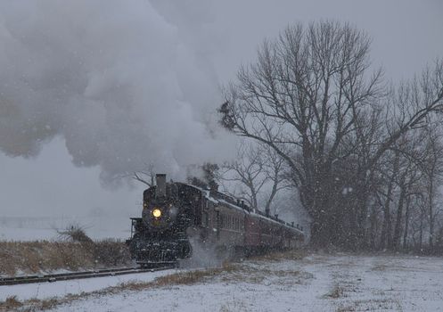 View of An Antique Restored Steam Locomotive Blowing Smoke and Steam Traveling Thru Farmlands and Countryside in a Snow Storm