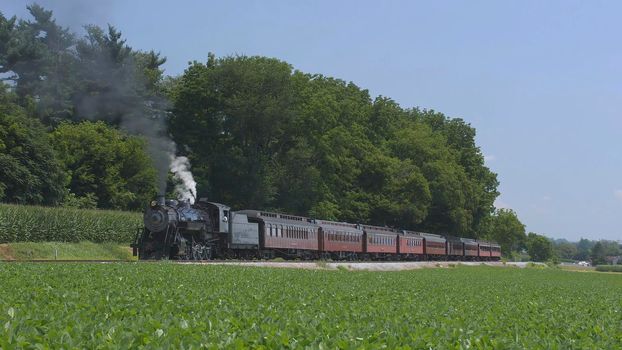 View of An Antique Restored Steam Locomotive Blowing Smoke and Steam Traveling Thru Farmlands and Countryside on a Sunny Summer Day
