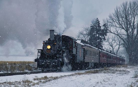 View of An Antique Restored Steam Locomotive Blowing Smoke and Steam Traveling Thru Farmlands and Countryside in a Snow Storm