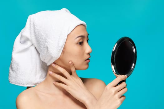 woman of asian appearance with bare shoulders looks mirror with a towel on her head blue background. High quality photo