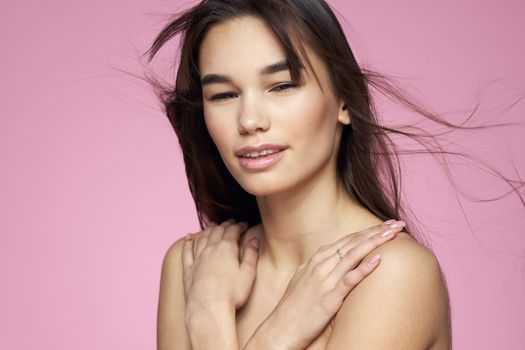 Brunette with bare shoulders holding her hair clean skin pink background. High quality photo