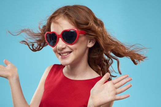 Little girl in sunglasses in the form of hearts red dress curly hair blue background. High quality photo