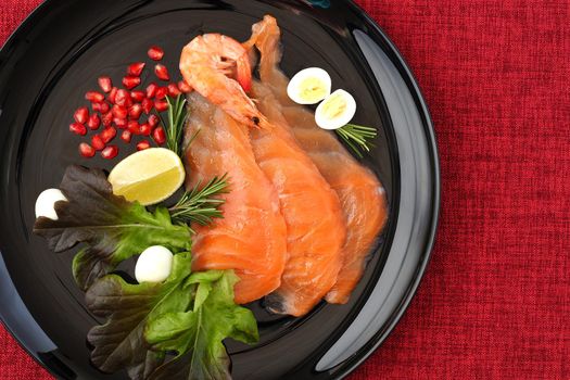 A seafood dish. Sliced salmon slices on a beautiful black plate with lemon, shrimp, quail egg and herbs and a red background. High quality photo