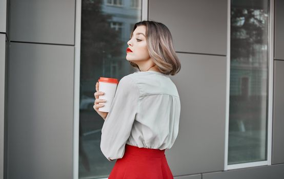 pretty blonde with a cup of coffee near the building on the street and red skirt white shirt. High quality photo