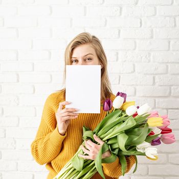 Mothers Day, Women's Day concept. Spring holidays. Happy beautiful woman in yellow clothes holding bouquet of tulips and blank card for mock up design