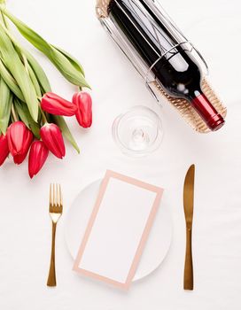 Mock up menu frame in restaurant or cafe. Top view of table setting with menu card, cutlery, fresh red tulips and wine on white fabric tablecloth