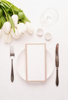 Mock up menu card in restaurant or cafe. Top view of table setting with menu card, cutlery, fresh white tulips, wine glass and candles for romantic dinner on white fabric tablecloth
