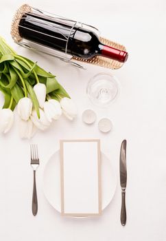 Mock up menu card in restaurant or cafe. Top view of table setting with menu card, cutlery, fresh white tulips, wine and candles for romantic dinner on white fabric tablecloth