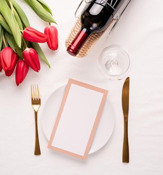 Mock up menu frame in restaurant or cafe. Top view of table setting with menu card, cutlery, fresh red tulips and wine on white fabric tablecloth