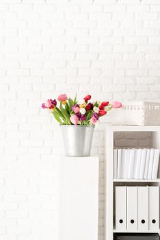 Bucket of fresh tulip flowers next to the bookshelf over white brick wall background