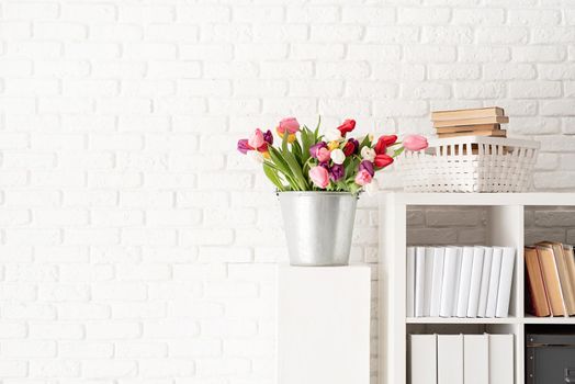 Bucket of fresh tulip flowers next to the bookshelf over white brick wall background