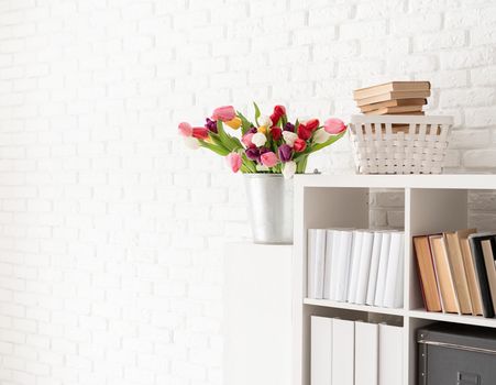 Bucket of fresh tulip flowers next to the bookshelf over white brick wall background