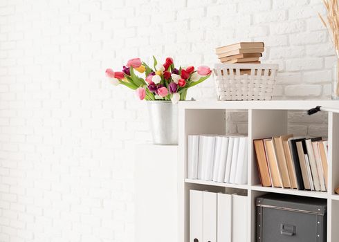 Bucket of fresh tulip flowers next to the bookshelf over white brick wall background